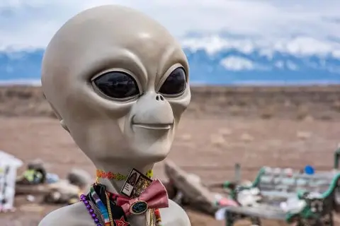 A statue of an alien stands outdoors with mountains in the background.