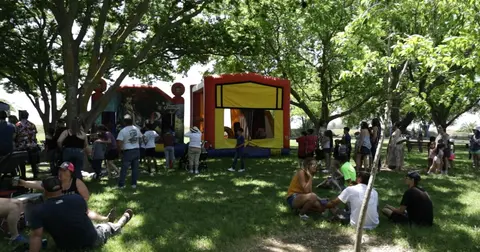 People gather in a park near bounce houses under shady trees.