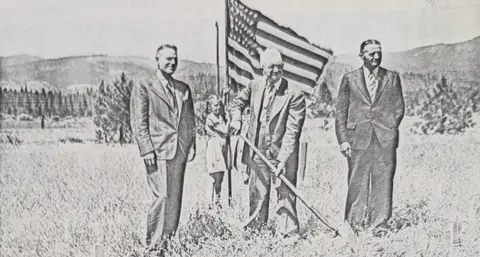 A historic black and white photo shows people with an American flag outdoors during a groundbreaking.