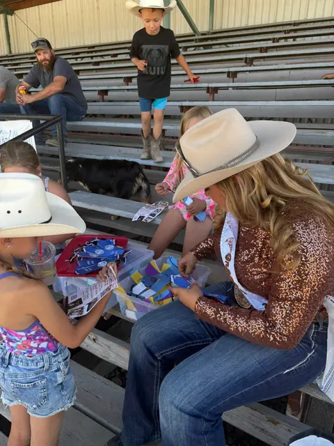 A woman in a cowboy hat and sparkling shirt sits in bleachers looking through ribbons with children.