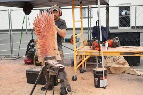 A man carves a wooden wing sculpture outdoors.