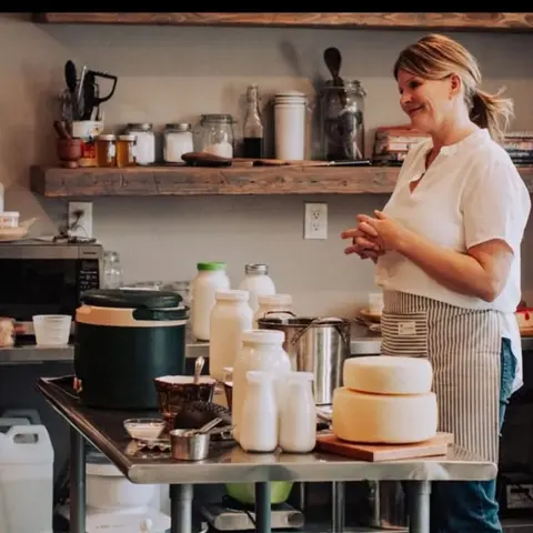 A woman stands in a kitchen next to a table with jars of milk and wheels of cheese.