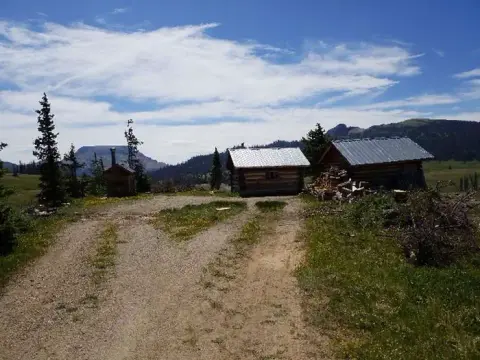 Small wooden cabins stand in a mountainous landscape under a cloudy blue sky.