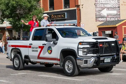 A white pickup truck participates in a street parade with people waving from the back.