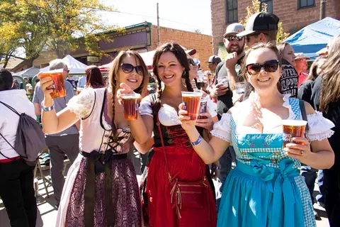 Three women in dirndls hold drinks outdoors at a festival.