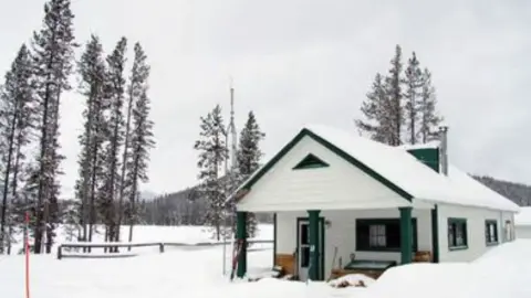 A white cabin with green trim sits in a snowy landscape with tall pine trees.