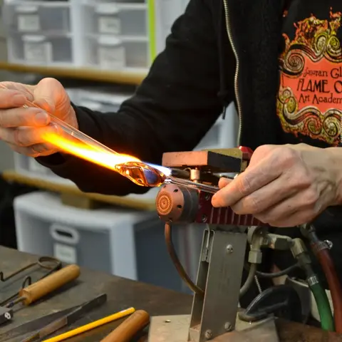 A person works with a torch to shape glowing glass.