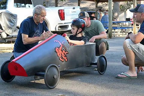 A child sits in a homemade soapbox car while adults look on.