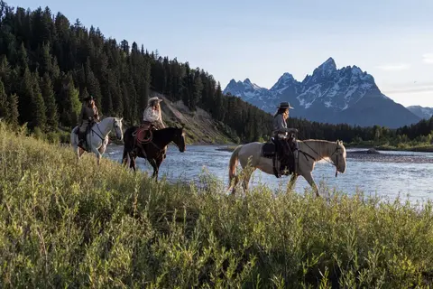Three people ride horses along a river with mountains in the background.