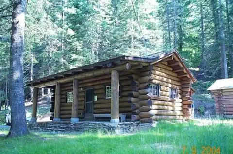 A log cabin sits in a forest clearing surrounded by tall trees.