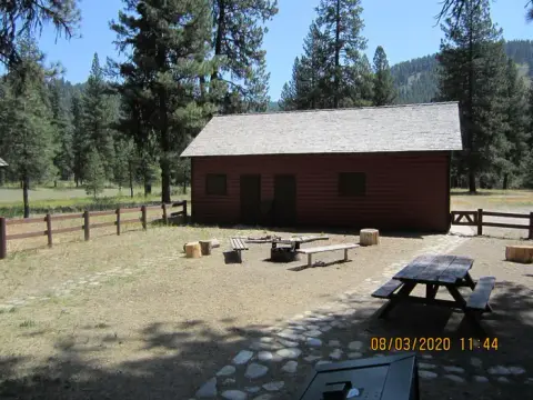A rustic wooden cabin with a campfire ring and picnic tables in a forested area.