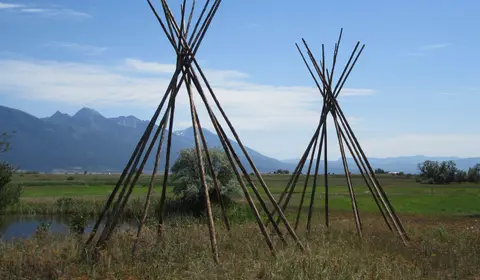 Two wooden teepee frames stand in a grassy field with mountains in the background.