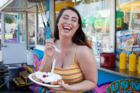 A smiling woman holds a plate of food in front of a food truck.