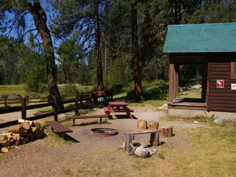 A wooden cabin with a green roof stands near a picnic area and fire ring in a forest.