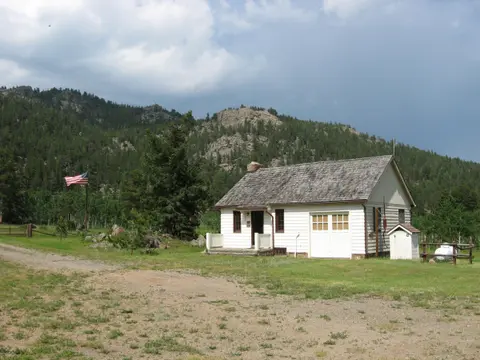 A white house with a garage and a flag pole sits in a grassy area with mountains in the background.
