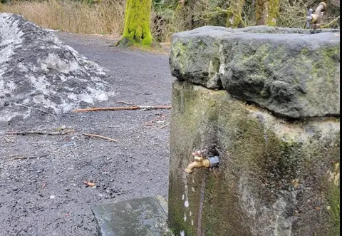 A stone water fountain stands outdoors with a running tap next to a pile of snow.