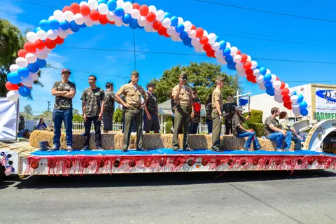 A decorated parade float carries people standing and sitting on hay bales beneath a balloon arch.