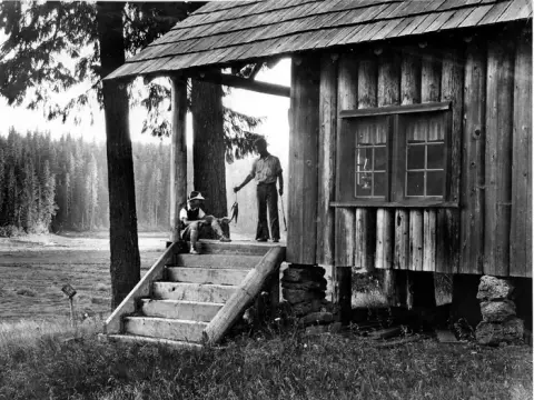 A historic black and white photo shows two people outside a rustic log cabin near a forest and water.