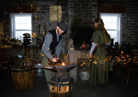 A blacksmith hammers glowing metal on an anvil as sparks fly in a workshop.