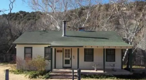 A single-story house with a green roof and a covered front porch, surrounded by bare trees and hills.