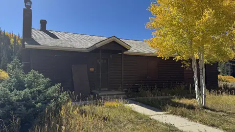 A dark wooden cabin with a covered porch stands amidst trees with yellow autumn leaves under a clear blue sky.