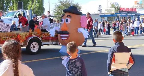 A large potato-shaped mascot walks along a street during a parade with onlookers watching.