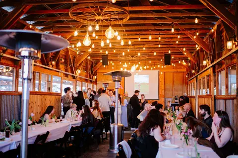 People gather at tables inside a rustic wooden venue decorated with string lights.