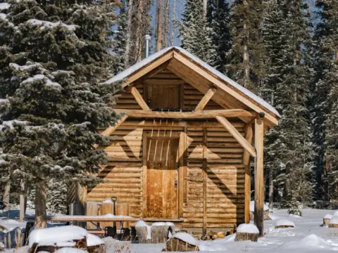 A wooden cabin in a snowy forest surrounded by pine trees.