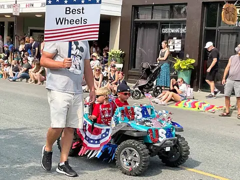 A man carrying a sign walks alongside two children riding in a decorated toy vehicle in a street parade.