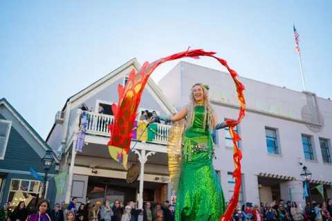 A performer in a green mermaid costume and red ribbons participates in an outdoor parade.