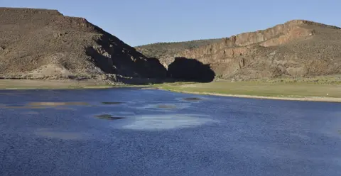 A river flows between dry, hilly terrain under a clear sky.