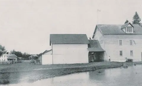 A historic black and white photo shows wooden buildings near a body of water.