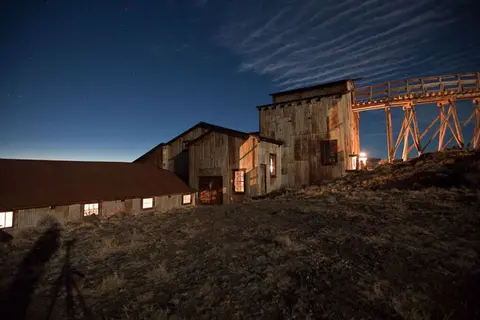 An old exterior building at night with a lit interior and a wooden bridge structure extending to the right.