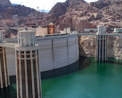 A large concrete dam stands in front of rocky hills with green water below.