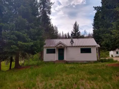 A small white cabin stands surrounded by trees and grassy terrain.