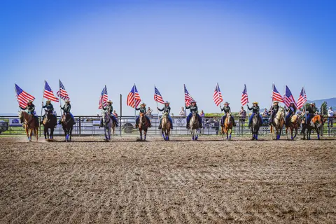 A line of horse riders holding American flags in a dirt arena.