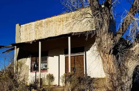 Exterior view of a weathered old building with a covered porch and a large tree in the foreground.