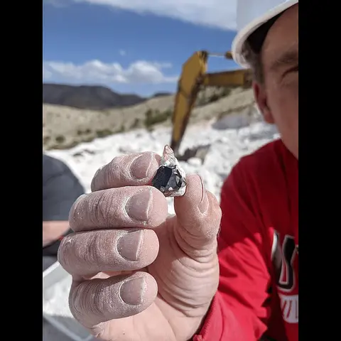 A person holding up a mineral crystal outdoors with an excavator in the background.