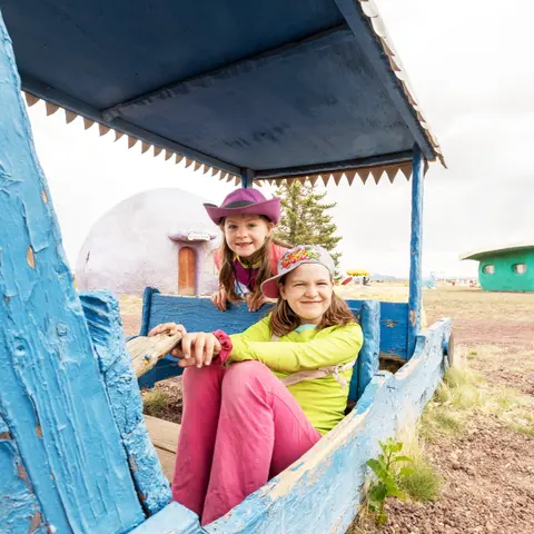 Two children sitting in a wooden toy vehicle outdoors.