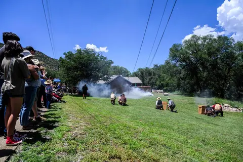 A crowd of spectators watches a historical reenactment outdoors on a grassy field.