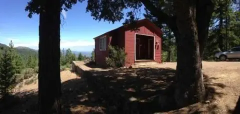 A red cabin stands outdoors among trees with a mountainous background.