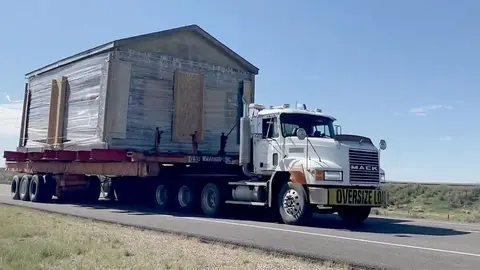 A large white semi-truck transports a small house on a flatbed trailer down a highway.