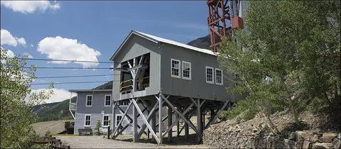 An old grey industrial building with wooden support stilts and a tall red structure behind it.