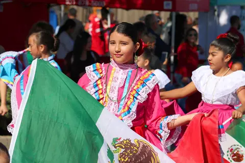 Young girls in traditional cultural dresses hold flags at an outdoor event.