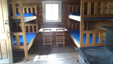 The interior of a rustic cabin features wooden bunk beds with blue mattresses and a table with chairs.