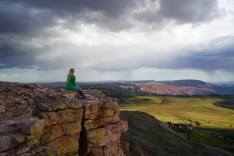 A person sits on a rocky cliff overlooking a vast landscape under a cloudy sky.