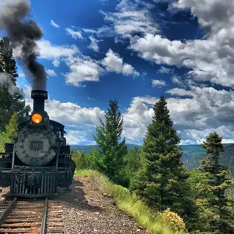 A steam train travels along a mountain railway line under a cloudy blue sky.