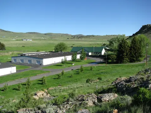 An elevated view shows buildings situated in a green valley surrounded by rolling hills and clear blue skies.