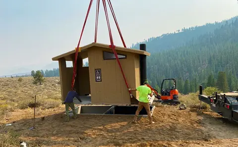 A prefabricated restroom building being lowered into place by a crane.