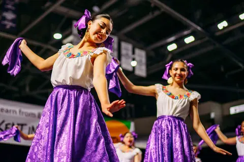 Dancers perform in traditional attire with purple skirts and hair ribbons.